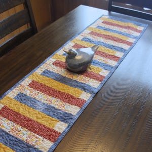 May include: A quilted table runner with a patchwork design in shades of blue, yellow, and red, with a small floral pattern. A ceramic bird figurine sits on the runner, adding a decorative touch to the table setting.
