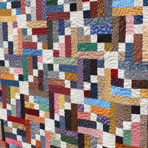 May include: A handmade quilt with a patchwork design. The quilt features a variety of fabric patterns and colors, including red, blue, green, and brown. The quilt is made of rectangular blocks arranged in a grid pattern, with white spaces between the blocks.