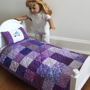 May include: A white wooden doll bed with a purple patchwork quilt and pillow. The quilt features various shades of purple squares. The pillow has a white center with embroidered flowers. The bed frame has a curved headboard and footboard.