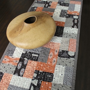 May include: A rectangular quilted table runner featuring a patchwork pattern of orange, gray, black, and white fabric squares with various geometric and text prints, topped with a smooth, round, light brown wooden bowl with a central opening.