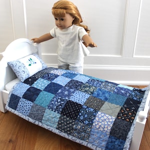 May include: A white doll bed with a blue patchwork quilt and matching pillow. The quilt features various shades of blue fabrics in a square pattern. The pillow has a floral design. The bed frame is white.