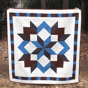May include: A handmade quilt with a central starburst design in shades of blue, brown, and white. The star is surrounded by a white background and framed by a border of alternating blue and brown squares. The quilt is displayed outdoors.