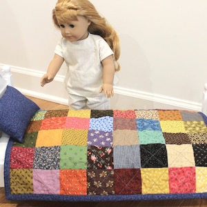 May include: A doll-sized white wooden bed frame featuring a patchwork quilt made of multi-colored fabric squares in various patterns including floral, geometric, and solid prints. A dark blue pillow rests at the head of the bed on the quilt.