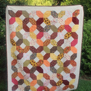 May include: A rectangular quilt featuring a repeating geometric pattern of octagonal shapes. The design is composed of various fabric patches in shades of yellow, orange, red, brown, and green, set against a light cream background with a brown border.
