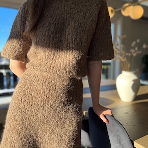 May include: A brown knitted two-piece outfit. The top is a short-sleeved crew neck jumper. The skirt is a mini skirt. The set is made of a textured, fuzzy material. The outfit is suitable for casual wear.