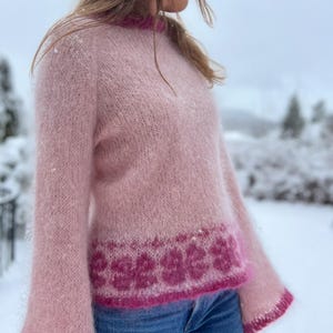 Pode incluir: Um suéter de malha felpuda rosa claro com mangas compridas e gola redonda. A peça apresenta um padrão floral rosa escuro ao redor da bainha e dos punhos, com acabamento rosa escuro correspondente ao longo da gola e das mangas.