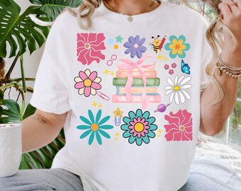 May include: White t-shirt with a colorful floral and book design. The design includes pink, yellow, blue, and teal flowers, a stack of books tied with a pink bow, and a cartoon bee with a pencil.