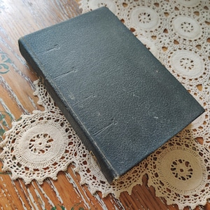 May include: A vintage, dark teal book with a textured cover. The book is resting on a cream-colored, crocheted doily, placed on a distressed wooden surface. The book's spine shows signs of wear.