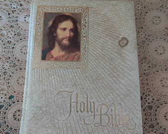 May include: A cream-colored Holy Bible with gold lettering and a portrait of Jesus Christ. The cover features a portrait of Jesus in a gold frame. The title "Holy Bible" is written in elegant gold script.
