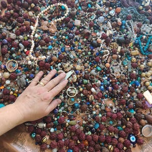 May include: A large assortment of jewelry components and beads in a copper-colored tray. The collection includes various sizes and colors of beads, necklaces, and pendants. A hand is resting on the assortment. The beads are in shades of brown, blue, white, and gold.