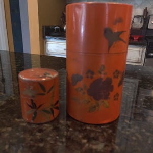 May include: Two orange cylindrical containers with floral and bird designs. The larger container has a bird silhouette and floral patterns in brown and gold. The smaller container has a lid and similar floral designs. Both containers are on a dark surface.