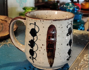 May include: A speckled beige ceramic mug with a brown and black design. The mug has a rounded shape and a handle.
