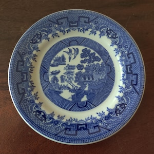 May include: A round, white and blue ceramic plate with a detailed Willow pattern design. The plate features a central landscape scene surrounded by floral and geometric borders. The plate is approximately 9 inches in diameter.