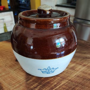 May include: A brown and white ceramic jar with a lid. The jar has a rounded shape with a brown top and a white bottom. A blue crown design is visible on the white section. The jar is sitting on a wooden surface.