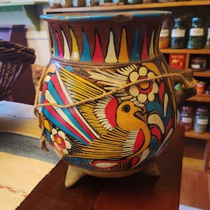 May include: A round, three-legged clay pot featuring a brown base with hand-painted designs of a yellow bird, white flowers, and colorful geometric patterns in red, blue, and yellow. A thin, light brown twine is wrapped around the pot's neck.