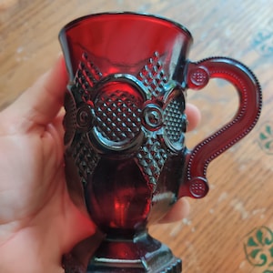 May include: A ruby red glass mug with a decorative pattern. The mug features a handle and a detailed design around the body, including diamond and floral shapes. The base is square and the mug is held in a hand.