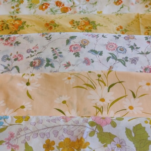 May include: A collection of folded fabric pieces featuring various floral patterns. The textiles display diverse botanical prints in shades of yellow, orange, pink, blue, and green on white and light peach backgrounds, showcasing daisies and wildflowers.