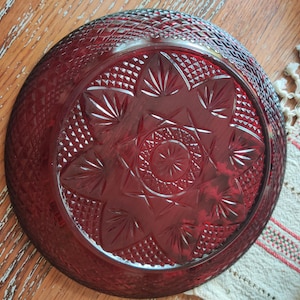 May include: A ruby red glass plate with a detailed starburst and diamond pattern. The plate has a wide rim and a central design of radiating lines and shapes. The plate is on a wooden surface.