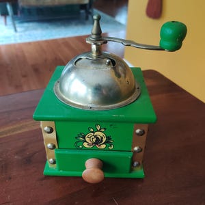 May include: A vintage coffee grinder with a green wooden base and a silver metal top. The grinder features a hand crank with a green wooden handle and a small drawer with a wooden knob. The front of the grinder has a floral design.