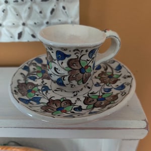 May include: A small, white ceramic teacup and saucer set. The cup and saucer are decorated with a floral pattern in brown, blue, and green. The set is placed on a white shelf, with a decorative white wall piece in the background.