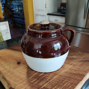 May include: A brown and white ceramic crock with a lid and handle. The crock has a glossy finish and sits on a wooden cutting board. The lid has a small, round knob.