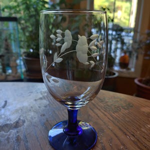 May include: A clear wine glass with a blue stem and base. The glass features an etched floral design. The glass is sitting on a wooden surface. The background is blurred, showing plants and a window.