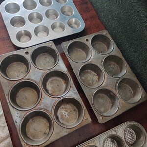 May include: A collection of vintage metal muffin tins in various sizes. The tins are made of aluminum and show signs of wear, with some having a darker, aged appearance. The arrangement includes tins with 6, 12, and 10 muffin cups.