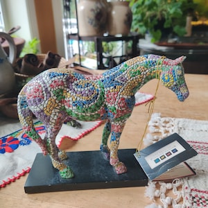 May include: A horse figurine featuring a colorful mosaic pattern of red, green, blue, yellow, and purple tiles. The horse stands on a rectangular black base and has a gold string attached to its neck with a tag labeled The Trail of Painted Ponies.