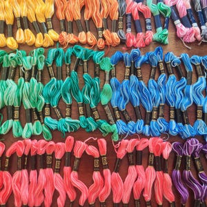May include: A collection of colorful embroidery floss in various shades, including yellow, orange, green, blue, pink, and purple. Each skein is neatly bundled with a label, ready for crafting projects. The floss is arranged on a wooden surface.