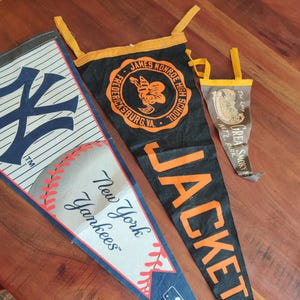 May include: Three vintage pennants on a wooden surface. One is a New York Yankees pennant with a baseball design. Another is black with orange text that reads "JACKETS" and a school emblem. The smallest is brown with text.