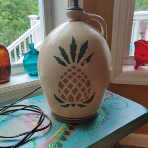 May include: Cream-colored ceramic lamp with a pineapple design in green and brown. The lamp has a handle and a black cord. The lamp is sitting on a teal table with floral designs.