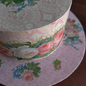 May include: A round, pink decorative box with a floral design. The lid and base feature a repeating pattern of pink flowers and a detailed floral border. The box is likely used for storage or decoration.