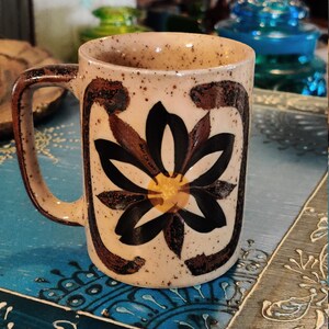 May include: A brown ceramic mug with a speckled glaze and a floral design. The mug has a brown handle and a white background with a black and brown flower with a yellow center.
