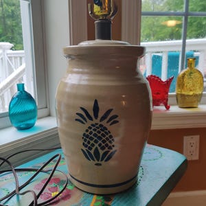 May include: A tan ceramic lamp with a blue pineapple design. The lamp has a black base and a gold-colored light socket. The lamp is sitting on a painted table with a floral design.