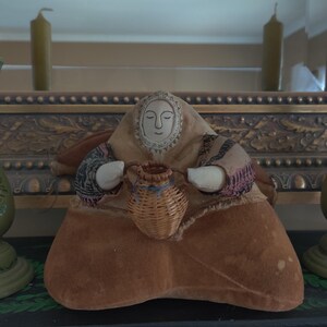 May include: A handcrafted doll with a serene expression, wearing a tan fabric outfit and holding a small woven basket. The doll's face is detailed with delicate features. The overall design suggests a folk art or vintage aesthetic.
