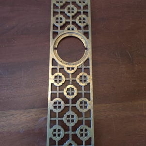 May include: A rectangular, brass-colored decorative door plate with an openwork design. The plate features a large central circular opening and a repeating pattern of stylized floral shapes. The metal has a slightly aged appearance.