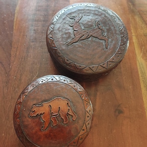 May include: Two round, dark brown wooden boxes with carved animal designs. One box features a leaping deer, the other a bear. Both boxes have a decorative border. The boxes are on a wooden surface.
