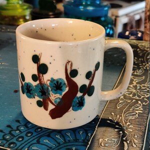 May include: White ceramic mug with a brown and blue floral design. The mug has a speckled glaze and a rounded handle.