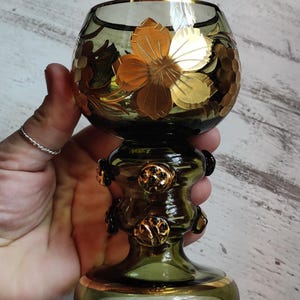May include: A green glass goblet with gold floral and grape vine designs. The goblet has a rounded bowl, a decorative stem, and a gold rim. The gold accents add a touch of elegance to the vintage glassware.