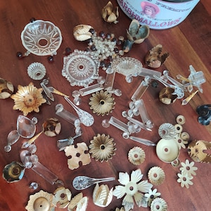 May include: A collection of vintage craft supplies, including clear glass crystals, metal flower-shaped embellishments, and small decorative pieces. A container of "Oxford's Premium Marshmallows" is visible in the background. The items are arranged on a wooden surface.