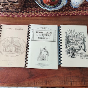 May include: Three vintage cookbooks with spiral binding are displayed on a wooden surface. The covers feature illustrations of a church, a town scene, and a church. Titles include "Yankee Sampler," "Home Town Recipes of Rockville," and "Out of Vermont Kitchens."
