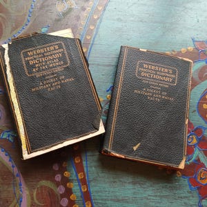 May include: Two vintage black dictionaries with gold lettering and trim. The books are titled "Webster's Practical Illustrated Dictionary" and include "A Digest of Military and Naval Facts."