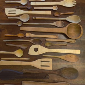 May include: A collection of various wooden kitchen utensils including spoons, spatulas, and tongs arranged on a flat surface. The assortment features different wood tones, shapes, and sizes, ranging from small tasting spoons to large ladles.