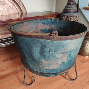 May include: An antique copper cauldron with a weathered, teal patina, resting on a wrought iron stand. The cauldron has a wide, open mouth and two small handles. The metal shows signs of age and use, adding to its vintage charm.