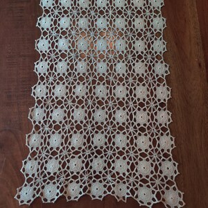 May include: Cream-colored, open-weave crochet table runner. The runner features a repeating pattern of circular motifs connected by delicate, star-shaped designs. The intricate lace-like texture is displayed against a dark wood surface.