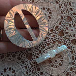 May include: A vintage, iridescent mother-of-pearl buckle and clasp set. The round buckle features a floral design around the outer edge. The clasp is rectangular with a similar iridescent sheen. The set is likely for a belt or other garment.