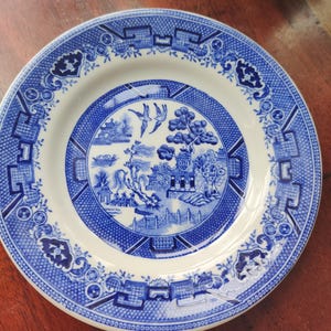 May include: A round, white and blue ceramic plate with a classic Willow pattern. The plate features a detailed blue landscape scene in the center, surrounded by a blue border with floral and geometric designs. The plate is approximately 10 inches in diameter.