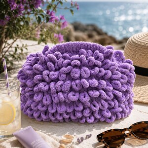 Könnte beinhalten: Eine violette Clutch mit einer texturierten Oberfläche aus geschlungenem Garn. Die Tasche hat eine abgerundete, voluminöse Form und besteht aus weichem, flauschigem Material. Sie liegt auf einer hellen Oberfläche neben einem Glas Limonade, einer Tube Sonnencreme und einer Sonnenbrille.