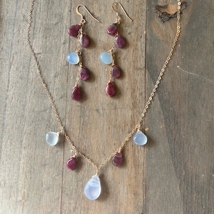 May include: A gold necklace and earrings set with a delicate chain and alternating light blue and burgundy gemstone drops.