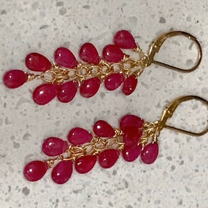 May include: A pair of gold-coloured earrings with a cascade of vibrant, teardrop-shaped red gemstones. The gemstones are strung together with delicate gold-coloured chains and wires, and secured with lever-back closures. The earrings are displayed against a light-coloured background.